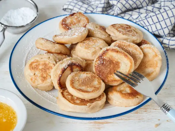Ein Teller voller goldbrauner Poffertjes, bestäubt mit Puderzucker. Ein Gabel liegt bereit, um das leckere Poffertjes Rezept zu genießen.