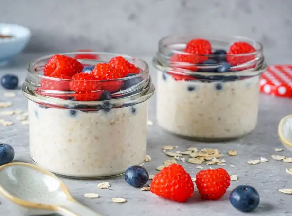 Zwei Gläser mit Overnight Oats, garniert mit frischen Himbeeren und Blaubeeren. Ein Löffel und Haferflocken liegen daneben.