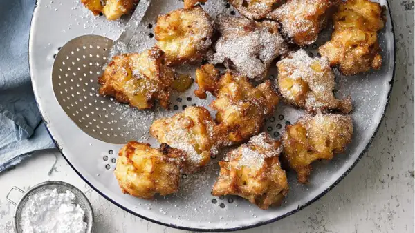 Goldbraune Apfelkrapfen, frisch aus der Pfanne, mit Puderzucker bestäubt, liegen auf einem Sieb.