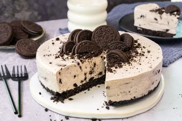 Ein köstlicher Oreo Kuchen, dekoriert mit ganzen und zerbrochenen Keksen, wartet darauf, angeschnitten zu werden. Daneben liegen Gabeln und ein Glas Milch.