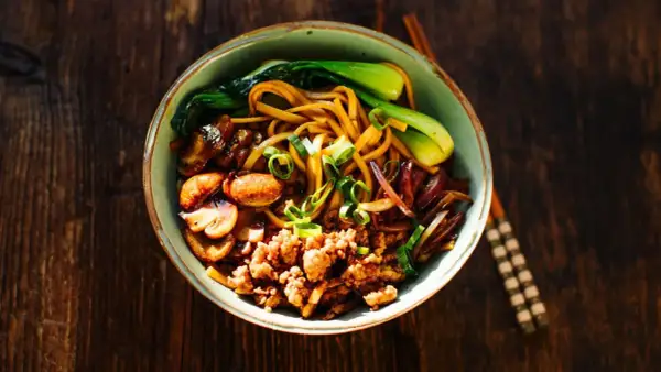 Herzhafte Udon Nudeln mit gebratenem Tofu, Pilzen, Pak Choi und Frühlingszwiebeln auf einem rustikalen Holztisch.