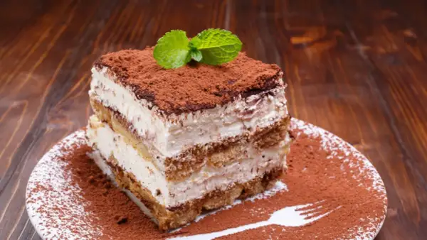 Ein Stück cremiges Tiramisu ohne Ei, bestäubt mit Kakao und garniert mit Minze, auf einem Teller mit Kakaopulver-Gabelabdruck.