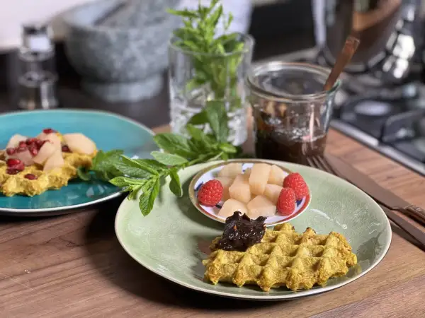 Zwei Teller mit goldgelben Haferflocken Waffeln, garniert mit Früchten und Marmelade, daneben frische Minze.