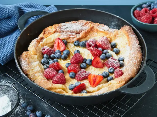 Ein goldbrauner Dutch Baby mit Puderzucker und frischen Beeren wie Erdbeeren, Himbeeren und Blaubeeren in einer Gusseisenpfanne.