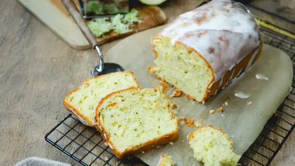 Saftiger Zucchini Kuchen mit Zuckerguss, frisch angeschnitten auf einem Gitter. Daneben liegen geriebene Zucchini und ein Hobel.