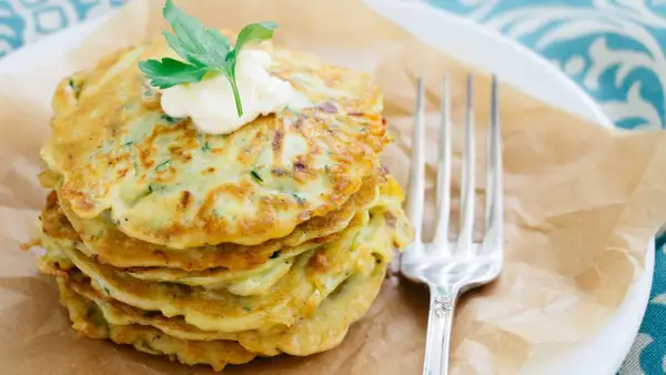 Ein Stapel goldbrauner Zucchini-Pancakes, garniert mit einem Klecks Sour Cream und Petersilie, daneben eine Gabel.