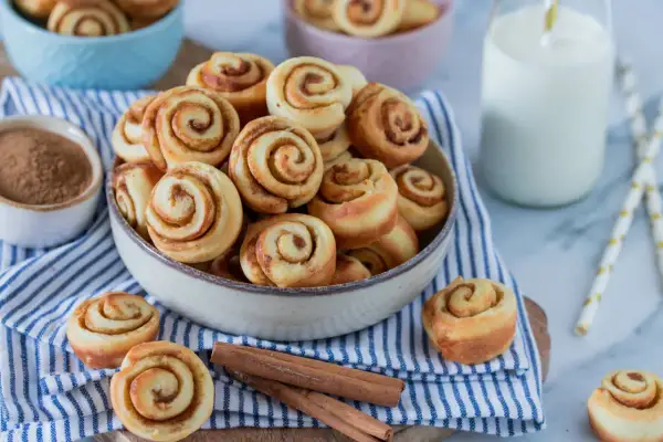 Mini Zimtschnecken in einer Schüssel, daneben Zimtstangen und ein Glas Milch.
