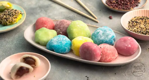 Bunte Mochi-Bällchen, die Lust aufs **Mochis selber machen** machen. Auf einem Teller liegen sie in verschiedenen Farben, daneben Schüsseln mit Sesam und roten Bohnen.