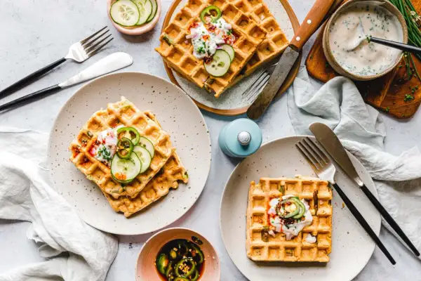 Drei Teller mit veganen Waffeln, garniert mit Gurkenscheiben, Zwiebeln und einer cremigen Sauce. Dazu ein Schälchen mit eingelegten Jalapeños.