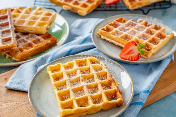 Goldbraune Waffeln, bestäubt mit Puderzucker, warten auf ein leckeres Waffeln Rezept. Eine Erdbeere garniert eine Waffel.