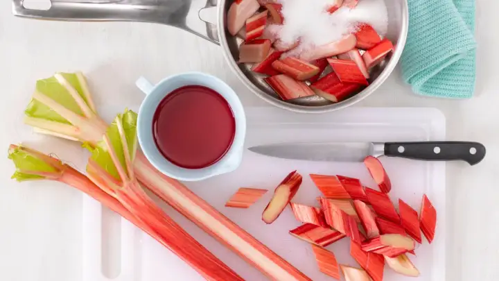 Frischer Rhabarber wird geschnitten und mit Zucker für Rhabarberkompott vorbereitet. Ein Messer und eine Schüssel mit rotem Saft liegen bereit.